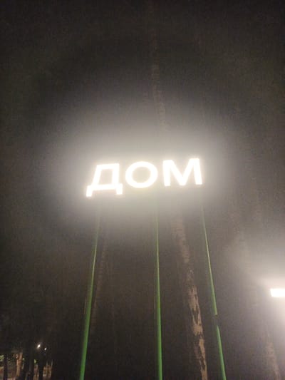 The image shows a neon sign that spells out "ДОМ" (DOM) in large, white, block letters, illuminated in the dark. The sign is mounted on tall, slender green poles. The letters are bright and emit a soft glow that slightly obscures the background. The surrounding environment is dark and appears to be a dimly lit outdoor setting, possibly a park or a street at night, with some trees visible in the background. There are also some distant lights in the lower left of the image, suggesting the presence of other structures or lighting. The overall atmosphere is somewhat mysterious due to the darkness and the bright, illuminated sign. The time of day is clearly night, and the weather appears to be foggy or hazy, contributing to the diffused glow of the neon sign and obscuring details of the background. There is no specific activity or event taking place, as the image focuses on the static sign and its immediate surroundings. The location context provided is Radkevshchina, Russia.