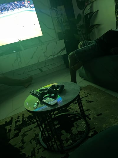 A person sits on a couch, feet resting on the floor beside a round, metal coffee table. The table holds two game controllers, remotes, and a small USB drive. A large television screen displays a bright scene, possibly a sporting event, with a crowd visible in the background. The floor is tiled with a marble-like pattern. A dark patterned rug is partially visible under the table, and a plant stands in the background.