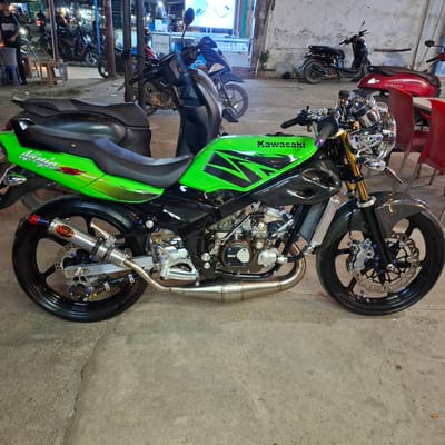 In Rhieng Blang, Indonesia, a vibrant green Kawasaki motorcycle, customized with carbon fiber accents and a polished exhaust, is parked on a cracked concrete surface. The motorcycle, a Kawasaki Ninja, is the central subject, with its prominent "Kawasaki" and "Ninja" branding visible. The scene appears to be an outdoor area, possibly a street or marketplace, indicated by the blurred background featuring other parked motorcycles and dimly lit stalls. The overall lighting suggests it is evening or night. The motorcycle is clean and appears to be in good condition, with custom blue bolts and a racing-style exhaust system. The surrounding environment is nondescript, with concrete ground and a weathered wall in the background.