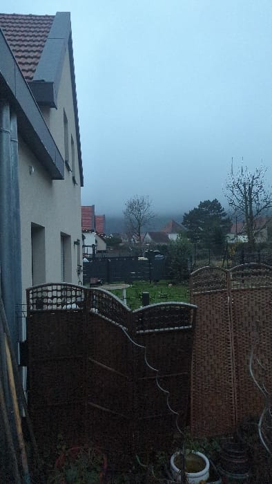 The image captures a static outdoor scene in a residential area of Châtenois, France, under overcast and hazy conditions. The light is soft and diffused, creating a muted atmosphere.
In the foreground on the left, a modern building with a dark gray tiled, sloped roof and light-colored walls is prominent. Several rectangular windows and a dark gutter system are visible on its facade.
To the right, a garden or patio area is depicted, featuring a tall, dark brown privacy screen or fence constructed from a woven material. In front of this, dark outdoor furniture, possibly chairs or a bench, with intricate decorative patterns, is arranged. A white pot with a yellow interior is visible near the fence.
The middle ground extends to a residential landscape with several trees, some bare of leaves, suggesting a non-summer season. Other houses with red-tiled roofs can be seen further back, partially obscured by the atmospheric haze that blankets the distant landscape.
The sky is a uniform light blue-gray, blending seamlessly with the foggy or misty horizon. The overall impression is one of quiet stillness on a dull, possibly chilly, daytime. No people or visible text are present in the image.