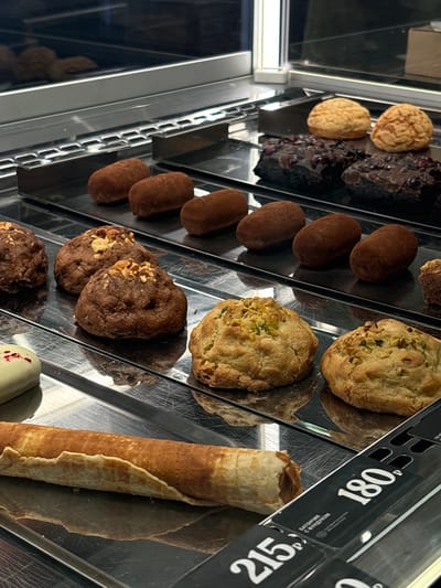 A display case in Borovoye Matyushino, Russia contains several types of desserts: round cookies, chocolate pastries, oval-shaped brown candies, pistachio cookies, chocolate cookies with nuts, and a rolled crepe. Price tags of 180 and 215 are visible on the right.