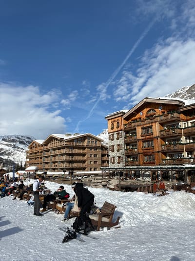 This image captures a vibrant daytime scene at a ski resort in Val-d'Isère, France. The foreground features a wide expanse of packed snow, functioning as an outdoor seating area. Several people, dressed in winter attire, are visible. Some are seated at wooden tables and chairs, many with cushions, enjoying the sunny weather. Others are standing, with skis and ski boots positioned nearby, indicating engagement in skiing activities.
In the mid-ground, a collection of grand, multi-story chalet-style buildings forms the backdrop. These structures boast traditional alpine architecture with prominent wooden facades, numerous windows, and balconies. Snow lightly dusts their roofs, blending them into the winter landscape. One particularly noticeable building features a stone base with wooden upper levels, and the word "AIRELES" is clearly visible on its ornate wooden gable. Another large wooden building stands behind it, equally grand.
The background is dominated by clear blue skies interspersed with scattered white clouds and a faint contrail. Distant snow-capped mountains complete the picturesque alpine setting. The overall impression is one of leisure and activity at a luxurious mountain destination, likely during midday given the bright, direct sunlight.