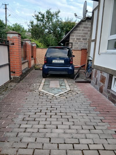 The image shows a narrow driveway or alleyway with a blue Smart car parked in the center, facing away from the viewer. The ground is paved with interlocking bricks, transitioning from red to gray in color. To the left, there is a brick wall and a gate. To the right, a building with a stone foundation and white siding is visible, with a blue bicycle leaning against it. Beyond the car, a smaller building or shed is partially visible, with a satellite dish mounted on its roof. Trees and foliage are present in the background, suggesting an outdoor, possibly residential, setting. The sky is overcast. The license plate of the car reads "AA 0570 OT". The location is identified as Vita-Poshtova, Ukraine.