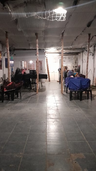 The image shows the interior of a building located at latitude: -34.667, longitude: -58.733 in the city of Merlo, Argentina. The room is sparsely furnished, featuring a large black piano covered in a red sheet. There is a row of chairs on the left, with another set of chairs on the right, covered in blue sheets. The room has tiled flooring and a high ceiling with a large, cracked, and discolored ceiling. The space appears to be undergoing renovation. There are exposed support beams and a large metal net hanging from the ceiling. The space appears to be an empty event space.  The building appears to be in a state of disrepair.