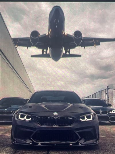 The image shows three dark-colored BMW cars parked in front of a building, with a commercial airplane taking off in the background. The cars are facing the viewer, with the closest car being a black BMW M2. The other two cars are partially visible behind it, also appearing to be BMW models. The airplane is silhouetted against a cloudy sky and is positioned above and behind the cars. The environment appears to be an airport or an industrial area, with a fence visible on the right side of the image. The scene is captured from a low angle, emphasizing the scale of the airplane and the cars.