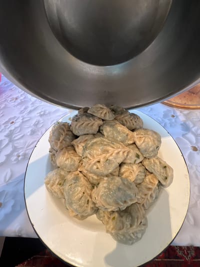 The image shows a close-up of a large white enamel plate filled with a pile of steamed dumplings. The dumplings are light-colored and have a pleated appearance, with some showing flecks of green herbs, likely chives or scallions, sprinkled on top. To the left of the plate is a large, dark-colored metal bowl, possibly a serving dish or a pot. Both the plate and the bowl are resting on a white tablecloth with a subtle floral embossed pattern. In the background, there are hints of other decorative elements, including a dark red textile with intricate patterns, suggesting a domestic setting. The lighting appears to be natural and soft, indicating it could be daytime. The scene is likely from Ak-Beshim, Kyrgyzstan, judging by the context provided. The focus is entirely on the food, suggesting the preparation or presentation of a meal. There is no visible text in the image.