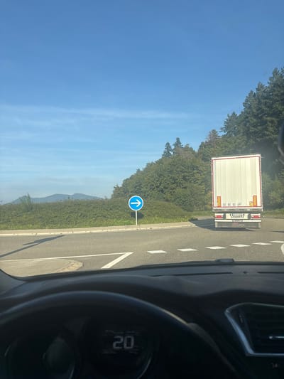 The image displays a road scene from a vehicle's driver-side perspective at a junction in Lièpvre, France. In the foreground, the upper section of a steering wheel and dashboard are visible, with a digital display showing "20," likely indicating speed. Ahead, the rear of a white semi-trailer truck occupies the right side of the frame, displaying "SCHMITZ CARGOBULL" and "LOCA" branding, along with speed limit stickers (60, 80, 90 km/h) and red and yellow reflective markings. To the left of the truck, a blue circular mandatory sign with a white right-pointing arrow is mounted on a pole, indicating required direction. The road surface ahead features white chevron and dashed line markings. Beyond the immediate road, a green, shrub-covered embankment transitions into a dense forest on a hillside to the right. In the background to the left, distant hills are visible beneath a clear blue sky with minimal cirrus clouds. No individuals are present in the visible scene.