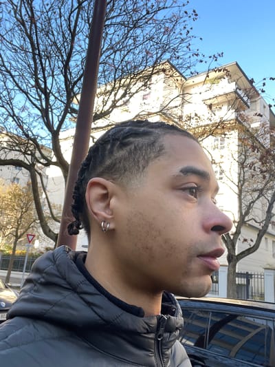 An individual, identified as a young male, is depicted in profile from the chest up, facing right, in Montigny-le-Bretonneux, France. He possesses dark skin, a short fade haircut on the sides, and dark cornrows styled on top. His left earlobe features two silver hoop earrings. Facial details include brown eyes, a defined nose, full lips, and a light stubble on the chin and jawline, with scattered minor epidermal blemishes on his cheek. He is attired in a dark, possibly grey or black, quilted puffer jacket over a dark, high-collared inner garment.
The background presents a clear, bright blue sky. Numerous deciduous trees, largely bare with sparse reddish-brown leaves, are visible, suggesting an autumn or winter setting. A tall, dark brown pole bisects the upper left portion of the frame. Behind the trees, multiple multi-story residential buildings with light-colored facades, flat or gently sloped grey roofs, and visible balconies are illuminated by sunlight. To the left, a portion of a red triangular 'yield' traffic sign is visible. In the lower right, a dark-colored vehicle reflects the surrounding environment. To the lower left, the front section of a lighter-colored vehicle is partially visible, displaying the initial characters "80" of a license plate. The overall scene indicates an urban outdoor environment under daylight conditions.