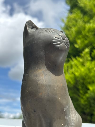 A close-up, low-angle, eye-level shot shows a weathered, dark grey, metal cat figurine. The figurine is seated, facing to the right of the frame, with its head tilted slightly upwards and to the right. Intricate lines suggest whiskers emanating from its muzzle, and its eyes are indicated by faint circular indentations. The cat's fur is suggested by subtle etched lines along its body.
The figurine is positioned outdoors, with a bright blue sky filled with fluffy white clouds forming the background. To the right of the figurine, a vibrant green, out-of-focus conifer tree dominates the background. The surface on which the figurine rests is not fully visible but appears to be white and flat, possibly a wall or ledge. The lighting suggests a sunny day, casting a soft shadow on the left side of the cat's body. The overall impression is one of a decorative object placed in a garden or outdoor setting. There are no people or animals present in the image. This scene does not contain any identifiable landmarks or features that would place it specifically in Oldham, United Kingdom.