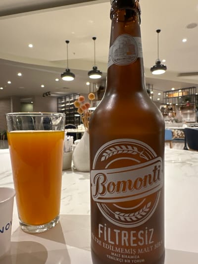 The image displays a brown glass bottle of "Bomonti Filtresiz" (unfiltered malt beer) exhibiting condensation, positioned prominently on a light-colored, possibly marbled table surface. The bottle's main label features the "Bomonti" logo with a wheat graphic, and text identifying it as "FİLTRESİZ FİLTRE EDİLMEMİŞ MALT BİRA." A secondary label near the bottle neck shows an architectural illustration. To the left, a clear glass contains an orange-colored liquid, approximately two-thirds full. A partial view of a white paper cup is visible to the extreme left. In the background, a modern interior space, likely a restaurant or hotel dining area, is evident. This setting includes multiple recessed ceiling lights and hanging black pendant lamps, a white decorative vase containing dried orange spherical flora, and various tables with blue chairs. Shelving units displaying bottles and other items are also present. The scene is artificially lit. The items are static, suggesting a pause in consumption. Based on the product shown, the location is consistent with Turkey, specifically in the city of Zeytinburnu. No individuals are present in the frame.