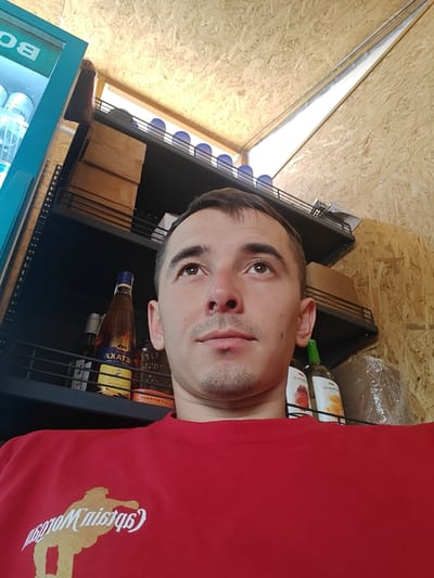 This is a close-up, eye-level, portrait-style photograph of a young man with short brown hair and a light beard, looking upwards. He is wearing a bright red t-shirt with a gold graphic design on it. The graphic appears to be a silhouette of a person with text above it. The text, which is partially obscured by the man's chin, seems to read "Captain Morgan".
The man is positioned in the foreground, taking up most of the frame. Behind him, there are shelves filled with various objects, including bottles of alcohol. One bottle is clearly visible with a yellow label that reads "AXATEM" in large, bold letters, and some smaller text below. Other bottles are present but less discernible. The shelves are black metal.
The background is made of light-colored, rough-textured wood paneling, suggesting an indoor setting. Above the shelves, there is a bright light source, possibly a skylight or an overhead lamp, illuminating the scene. To the left of the frame, part of a blue refrigerator or cooler is visible, with the word "B" in white letters on its side.
The lighting is bright, suggesting it could be daytime. The overall mood of the photo is casual and candid. There are no obvious indicators of a specific event or activity, other than the man posing for a selfie. The location context provided is Shkurupiivka, Ukraine.