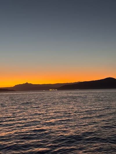 The image depicts a serene seascape at dusk, likely taken from a boat or the shore in Angra dos Reis, Brazil. The sky transitions from a deep, clear blue at the top to a vibrant orange hue near the horizon, indicating the sunset. Silhouetted against this colorful sky are several mountain ranges and hills. In the distance, lights from a town or settlement twinkle along the coastline. The foreground is dominated by the dark, rippling surface of the water, reflecting the warm tones of the sunset, creating streaks of orange and gold across the waves. The overall atmosphere is tranquil and picturesque, capturing the natural beauty of the Brazilian coast.