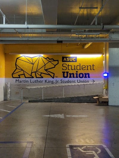 A bright yellow sign on a wall reads "ASUC Student Union" with a geometric bear design. Text below the sign says "Martin Luther King, Jr. Student Union" followed by an arrow pointing right. Adjacent to the wall is a gray concrete walkway with a blue line. A white arrow is painted on the walkway, pointing forward. Further down, a handicapped parking symbol is visible. The image is taken indoors, possibly in a parking structure or covered area. Metal piping and beams are visible above and to the left.