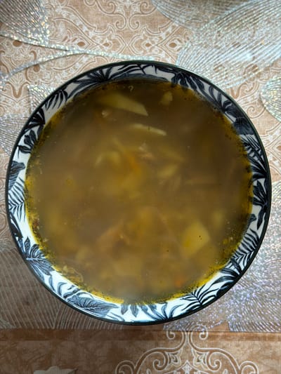 A bowl of soup sits on a patterned table in Заболотово, Russia. The soup is a murky brown color and appears to contain potatoes and possibly other vegetables. The bowl has a white, black, and blue leaf pattern. The table has a beige and brown patterned cloth with some silver accents.