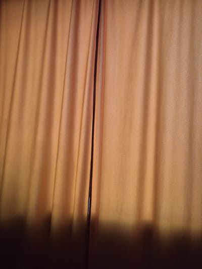 A curtain, predominantly orange-brown in color, hangs in vertical folds, creating a textured surface. A dark vertical line, likely the edge of another curtain or a structural element, bisects the image slightly to the right of center. The lower portion of the curtain is cast in shadow, transitioning from a deep brown to a lighter, muted tone. The background is not visible. The image was taken indoors.