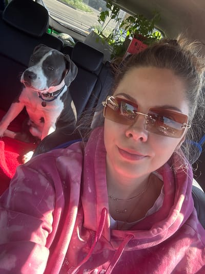 The image is a selfie taken from inside a car. A woman with her hair in a messy bun is wearing sunglasses and a pink tie-dye hoodie. She has two necklaces. A gray and white dog lies on a red blanket on the back seat of the car. A plant sits on the rear seat, likely a gift or a purchase to be transported. Outside the car, a glimpse of trees, buildings, and a road suggests an urban environment, though no specific location can be determined without further information, despite the request for Zavorovo, Russia.