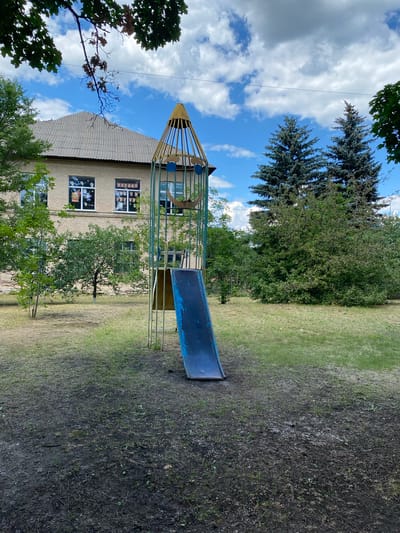 In the city of Markivka, Ukraine, on a cloudy day, a blue metal slide is part of a rocket-shaped playground structure. The slide extends from a metal frame with blue circular decorations and a yellow conical top, set against a backdrop of a brick building with a grey tiled roof. Two mature evergreen trees stand behind the building, with dense green foliage surrounding the playground. The ground is a mix of patchy grass and dark soil, with some dry leaves scattered across the area. Overhead, a wire stretches across the frame.