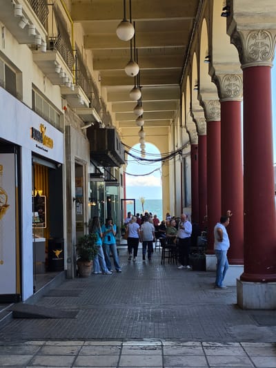 The image depicts a view along a colonnaded walkway with a row of large, dark red columns supporting an arched ceiling. The walkway is paved with gray tiles, and a few people are visible walking or standing. On the left side of the image, there is a gelato shop named "BUFALA GELATO" with a bright yellow interior. Further down the walkway, tables and chairs are set up, suggesting an outdoor seating area for a cafe or restaurant. In the background, the sea is visible under a bright sky. The architecture, particularly the columns and arches, suggests a historical or neoclassical style, common in cities like Thessaloniki, Greece. The scene appears to be a public space, likely a promenade or street, where people are enjoying a day out by the sea. The lighting suggests it is daytime, with sunlight casting shadows on the ground.