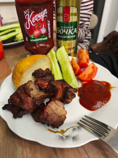 A plate of food is centered on a wooden table, consisting of several pieces of grilled meat wrapped in bacon, a slice of bread, cucumber sticks, and tomato wedges. A dollop of ketchup is placed to the side of the food, and a fork rests on the edge of the plate. In the background, a bottle of ketchup and a bottle of beer are visible, along with a person wearing a striped shirt and a dog. The scene appears to be indoors, likely in a home setting, and there is no indication of any specific location within the city of Речицы, Russia.