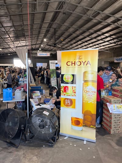 The image depicts a crowded indoor market area. Located at -27.44 latitude and 153.072 longitude, this is the Hamilton Indoor Markets in Hamilton, Australia.  A large poster for Choya Japanese liqueur dominates the right side of the image. The poster features images of the beverage,  with text in Japanese and English. Two men are standing near the poster, one wearing a blue shirt and the other a black shirt.  A woman with long dark hair is standing behind them, facing the camera, looking to her right.  To the left of the poster are two large industrial-style fans, while tables, chairs, and a diverse assortment of boxes and packaging material are scattered around.  In the background are numerous people, mostly blurred, with a few individuals in focus.  The scene suggests a bustling market with attendees interacting, browsing, and sampling products. The environment appears to be a large, open space with a high roof and metal trusses overhead.  A large white banner on the left side of the image is also visible, with the word "SAKE" visible in large letters.