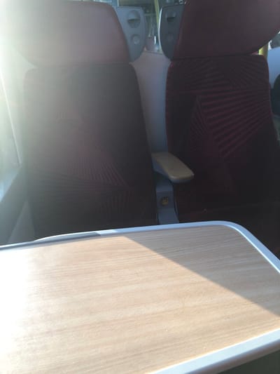 The image depicts the interior of a passenger transport vehicle, likely a train or bus. The specific location within Dettwiller, France, cannot be determined from the visual information provided.
In the foreground, a light-colored, wood-grain pattern tray table is visible, extending across the lower portion of the frame. Its left side is brightly illuminated by direct sunlight, which also causes significant lens flare in the upper left quadrant. The table features a light grey or white border.
Beyond the tray table, two high-backed passenger seats are positioned in a row. Both seats are upholstered in a dark red or maroon fabric with a subtle geometric pattern composed of radiating lines or stripes. The headrests are solid dark red. The left seat is largely obscured by intense sunlight and lens flare. The right seat shows more detail, including a beige-topped armrest with a grey base located between the two visible seats. Behind the headrests, light grey panels with circular elements, possibly controls or speaker grilles, are faintly discernible. No individuals are present in the scene, and no specific actions or interactions are depicted. The environment is clearly an interior space designed for passenger travel.