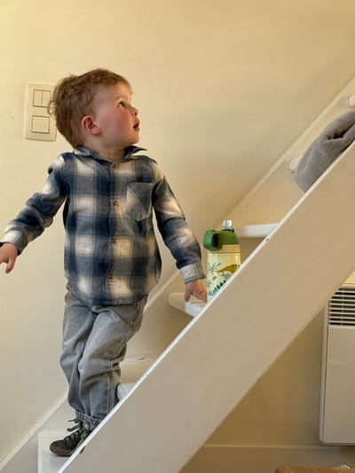 A young child, likely a toddler, is captured indoors, possibly near a staircase or a structural architectural element. The image is oriented sideways, depicting the child seemingly standing or interacting with a light-colored wooden banister or railing.
The child has light brown, curly hair and fair skin with rosy cheeks. They are wearing a long-sleeved plaid shirt in shades of blue, white, and tan, paired with grey pants or jeans, and dark athletic shoes with lighter laces. The child's head is tilted upwards, and they are looking towards the upper right of the frame with a curious or engaged expression.
In the background, a light beige or cream-colored wall is visible. A grey towel or fabric is draped over a part of the railing in the upper left corner. A green and yellow water bottle or sippy cup with a subtle floral or leafy pattern is tucked beneath the railing, near the child's right hand. Towards the bottom left, a set of white light switches or outlets is mounted on the wall. In the upper right background, partially obscured by the railing, there is a white rectangular vent or air conditioning unit. Another wooden surface is visible in the far upper right.
The scene suggests an ordinary daytime moment, possibly in a home, with the child exploring or playing. No specific time of day, weather conditions, or visible text can be definitively determined from the image itself, beyond the general indoor setting in Bredene, Belgium. The child's expression conveys curiosity and attentiveness.