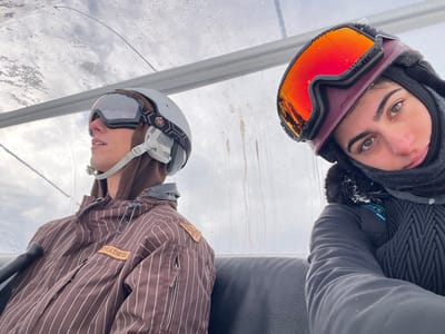 Two individuals are seated inside an enclosed ski lift cabin in Valtournenche, Italy, beneath an overcast sky. The transparent ceiling of the cabin exhibits numerous water droplets. On the left, a male subject is visible, wearing a light grey ski helmet with dark straps, silver-reflective ski goggles, a brown hooded sweatshirt, and a brown ski jacket featuring white vertical pinstripes and "FEATHERS" branding. He is looking upwards and to the right, away from the camera. On the right, a female subject is present, wearing a maroon ski helmet with orange-reflective ski goggles that display visible scratches or cracks, a black balaclava or neck warmer, and a dark grey textured ski jacket. Small flakes of snow or ice are present on her right shoulder. She is looking directly at the camera with a neutral expression. The seats of the cabin are dark in color.