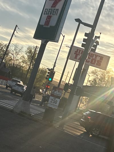 The image depicts a tilted, eye-level view of an urban intersection in Reno, Nevada, during what appears to be golden hour, characterized by strong sunlight from the right causing significant lens flare. Overhead, the sky is partly cloudy.

Prominently featured are two commercial signs: a large, green and red "7-ELEVEN" sign on a tall pole in the upper left, and a red and white "JIMBOY'S TACOS" sign further back and to the right. A traffic light assembly is central, showing a green signal for vehicular traffic, while an adjacent pedestrian countdown signal displays a red "14". Below this, a smaller street-side advertisement board is visible, promoting "$2.50 SAVE BY 2 MARLBORO PACKS".

At the intersection, a white pickup truck and a silver sedan are visible, both facing away from the camera, positioned on a street with a marked pedestrian crosswalk. A dark-colored SUV or crossover is partially visible in the lower right foreground, also facing away. A single pedestrian, dressed in light clothing, is observed walking from right to left across the crosswalk in the mid-ground.

The scene is populated with multiple utility poles and overhead power lines. Trees with varying degrees of foliage are present in the background, suggesting a seasonal transition or mixed species. The ground consists of asphalt road surfaces and concrete curbs and bases for the poles and signs.