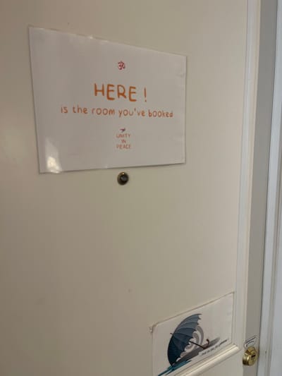 An interior, eye-level forensic view depicts a light-colored, possibly off-white, door with two affixed informational signs and a door peephole. The door is slightly ajar on the right, revealing a portion of a light-colored door frame and an adjacent space.
**Objects:**
1. **Top Sign:** A rectangular white sign, approximately A4 size, is positioned centrally on the upper half of the door. It features handwritten or drawn text in an orange-red color. The Om symbol (ॐ) is at the top center. Below it, the text reads: "HERE!" on one line, followed by "is the room you've booked" on the next. Below this, a small bird silhouette is present above the text "UNITY IN PEACE". The sign appears to be laminated or encased in a clear plastic sleeve.
2. **Door Peephole:** Directly below the top sign, a circular, metallic (likely brass or gold-toned) door peephole is installed.
3. **Bottom Sign:** A smaller, rectangular white sign is affixed to the lower half of the door. This sign displays a graphic of an open blue umbrella with raindrops falling and pooling water below it. Below the graphic, partially visible text in black reads: "...not in the flat please !". This sign also appears to be laminated or in a clear sleeve.
4. **Door Hardware:** On the right edge of the door, where it meets the frame, a brass-colored doorknob and a separate deadbolt lock mechanism are visible. A small, white sticker with partial text, possibly "OPEN unless," is affixed to the door frame immediately adjacent to the lock.
**Environment:** The scene is an indoor setting, likely a hallway or common area leading to a private room. No individuals are present. The precise location within Saint-Gilles - Sint-Gillis, Belgium, cannot be determined from the visual evidence.