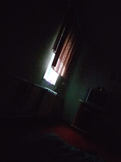 The image depicts a dimly lit room, possibly a bedroom, with a window on the left side. The window is partially covered by a patterned curtain. Natural light filters through the gap in the curtain, illuminating a portion of the room. A radiator is visible below the window. In the right side of the image, a television set with a glowing yellow light on top is placed on a wooden surface, likely a table or cabinet. The room appears to be a residential interior located in Verkhniy Mamon, Russia. The overall atmosphere is dark and somewhat austere. There are no visible individuals in the frame.