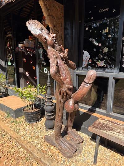 A wooden statue of a man is lying on a surface with his legs outstretched. The statue's head faces left, and the man is holding something in his hands. A large phallus extends upwards from the statue's groin. The statue is positioned next to a building with windows decorated with Christmas-themed stickers. A potted plant sits near the base of the statue. A wooden bench and gravel ground are also visible in the background.