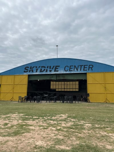 The image displays the exterior of an arched hangar building labeled "SKYDIVE CENTER" in dark lettering on its blue roof, located at an aerodrome in Chascomús, Argentina. The hangar features large yellow corrugated metal sliding doors, which are partially open, revealing a light aircraft inside. In front of the hangar, on a sparse grassy area with visible soil, approximately five black plastic tables are arranged with multiple accompanying black plastic chairs. To the right, near the open hangar section, a black speaker or audio equipment is positioned on the ground. Two individuals are partially visible inside the hangar opening on the right, near the speaker. The sky above is overcast with light grey clouds. A thin, dark pole extends vertically from the hangar roofline into the sky, where a small, dark, unidentified object is faintly visible.