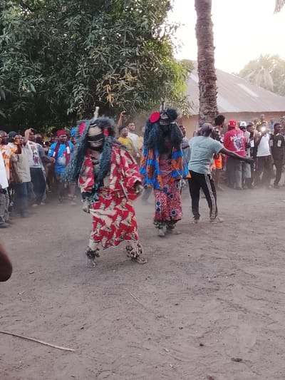 The image depicts an outdoor event in Tabu, Nigeria, on an unpaved, dusty ground. Two masked figures, identified as masquerades, occupy the central foreground. The left masquerade wears a full-body costume of red and white patterned fabric, grey shaggy material on the upper body, a dark striped mask, and natural fiber or shell rattles on its ankles, with dust visible around its feet indicating movement. The right masquerade wears a full-body costume of blue, red, and yellow patterned fabric, dark shaggy material, a dark mask with a red circular accent, and similar ankle rattles. An unmasked male individual, dressed in a light green t-shirt and black athletic pants, stands with arms outstretched between the right masquerade and the surrounding crowd. A dense crowd of predominantly male spectators, some observing and others holding mobile phones, forms a background. Vegetation includes a large leafy tree to the left and a tall palm tree to the right, with structures featuring corrugated metal roofs visible further back. A small, thin stick lies on the ground in the lower left.