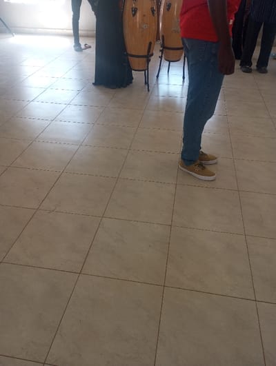 The scene is located indoors in Racecourse ward, Kenya, featuring a light beige tiled floor with visible grout lines. Four individuals are partially visible, standing. In the immediate right foreground, a person wears blue denim jeans and tan canvas shoes. To the left, two light brown conga drums on black metal stands are centrally positioned. Behind the drums, another person is seen wearing a long black garment. Further in the background to the left, the lower legs of a third person in dark trousers and shoes are visible, with a red sandal or slipper near their foot. The lower legs of a fourth person in dark attire are visible on the far right background. A bright light source from the left illuminates a pale wall in the background.