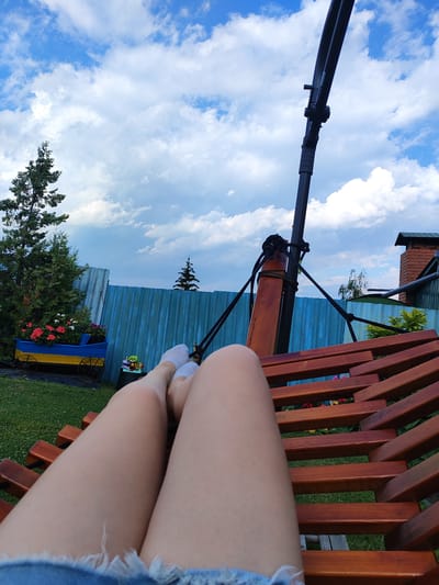A POV shot captures a person relaxing on a wooden swing. Their legs are extended forward, clad in white socks and denim shorts with frayed edges. The swing is suspended by a metal frame against a backdrop of a blue sky with scattered white clouds. To the left, a tall green evergreen tree stands near a blue fence adorned with a decorative cart filled with red and white flowers. The environment suggests a backyard in Krasnokutsk, Ukraine, with a hint of a brick chimney visible in the upper right corner.