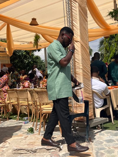 An outdoor social event is captured in Kumasi, Ghana, taking place under a large, translucent light orange canopy structure. The ground consists of uneven light-colored paving stones in the foreground, transitioning to artificial green turf and then more natural stone.
In the immediate foreground, a man with dark skin, short dark hair, and a trimmed beard walks from right to left, his head bowed slightly, looking downwards. He is dressed in a light green, short-sleeved tunic-style shirt, black trousers, and brown leather loafer-style shoes. He wears red-rimmed glasses and a dark watch on his left wrist. His right hand is raised to his face, near his glasses, while his left hand holds a clear plastic water bottle that appears to be supporting a dark-colored rectangular object, possibly a smartphone. An empty plastic water bottle and a green leaf lie on the paving stones near his feet.
To the man's right, a pillar wrapped in pleated light orange fabric stands, from which metal chains are suspended. A dark-colored plastic stool or small table is positioned near the pillar.
In the mid-ground to the left, several round tables are arranged, each covered with a white tablecloth and decorated with floral centerpieces featuring pink and red flowers, along with glassware and other bottles. Woven-backed chairs are placed around these tables. Multiple individuals, predominantly women dressed in colorful patterned attire, are visible standing and sitting around these tables. Above, the canopy is adorned with woven basket-like light fixtures and green foliage.
To the right in the mid-ground, other guests, primarily men, are seated at similar tables. Some are wearing garments matching the green hue of the main subject's shirt. The background reveals lush green trees and foliage under a bright sky. The scene depicts a daytime gathering, likely a reception or celebration.