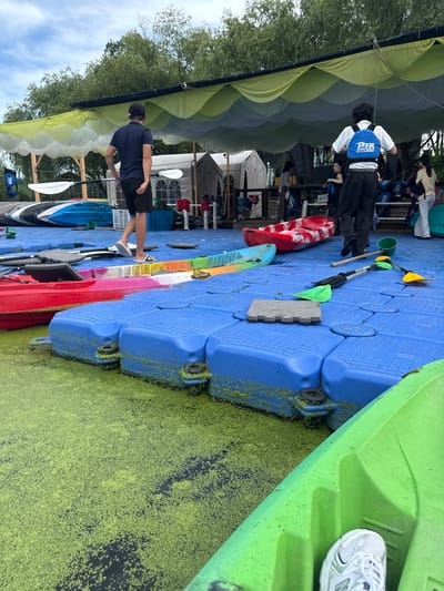The image depicts an outdoor scene at a waterfront recreational facility, likely in Lin'an District, China. The foreground is dominated by a blue modular floating dock covered in a thick layer of green duckweed, with several kayaks and paddles scattered across its surface. A bright green kayak is partially visible in the lower right corner, with a person's white athletic shoe resting inside it.
In the midground, several people are present. A man in a dark blue polo shirt and black shorts, wearing a black baseball cap, stands on the dock holding a paddle. To his right, another person wearing a blue life vest with "PTB POWER TO ACHIEVE" printed on it walks away from the camera. In the background, a large, translucent yellow and white canopy provides shade over tents and other structures. Other individuals can be seen walking around and interacting near the tents. The overall environment suggests a kayaking or boating rental or activity center.