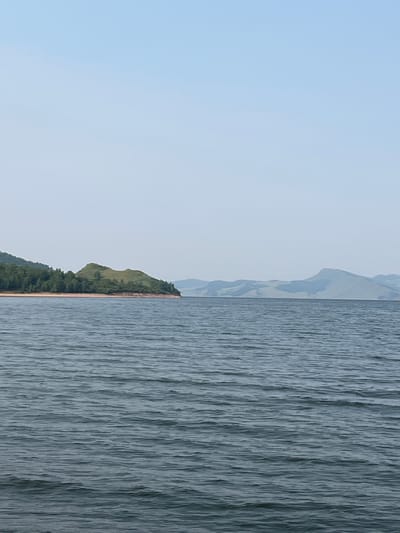 The image shows a wide expanse of calm, dark blue water in the foreground. In the middle ground, a green, rounded hill with a sparsely vegetated summit juts out into the water from the left. The hill is bordered by a sandy shore and a dense forest of green trees on its left flank. Further in the distance, a series of hazy, blue-green mountains form the horizon. The sky above is a clear, pale blue, with no clouds visible. The scene appears to be a natural landscape, possibly a lake or a wide river, near Novoselovo, Russia, given the context provided. No people or man-made objects are visible.