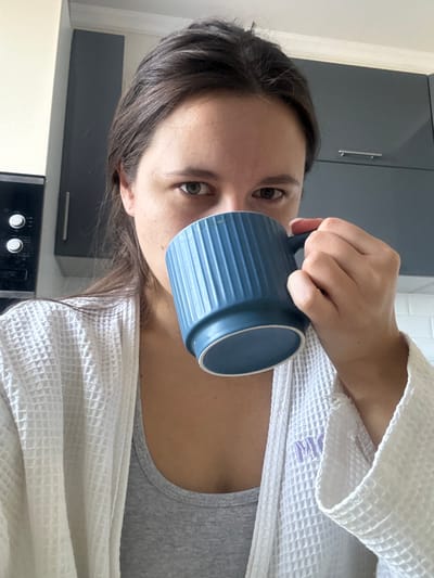 A frontal close-up image depicts a person, appearing female, in a kitchen in Селекционер-2 Снт, Russia. The individual, with dark brown hair, is visible from the chest up, looking directly at the camera. They are holding a teal blue, vertically ribbed ceramic mug with their right hand to their mouth, partially obscuring the lower half of their face. The person is wearing a white waffle-knit robe over a gray tank top; a light purple letter "M" is embroidered on the robe's left lapel area. The background features light-colored walls, dark grey overhead kitchen cabinets, a black appliance with two circular controls integrated into a cabinet on the left, and a white subway tile backsplash on the right.