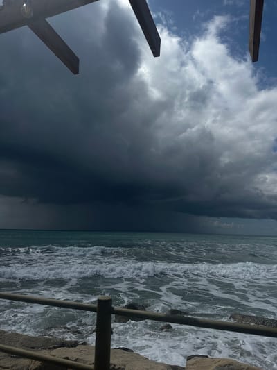 The scene depicts a coastal environment in Episkopi Lemesou, United Kingdom, under a developing storm. The upper frame is partially obscured by two dark wooden beams and a suspended light fixture. The sky is largely covered by dark, heavy stratocumulus clouds, with a concentrated area of rainfall visible over the distant ocean to the left. A lighter patch of blue sky and dispersed white clouds are present on the upper right. The sea is active, displaying numerous white-crested waves approaching a rocky or concrete embankment. A grey metal railing with a single visible post in the lower-left foreground separates the immediate viewing point from the shoreline. No individuals are present.