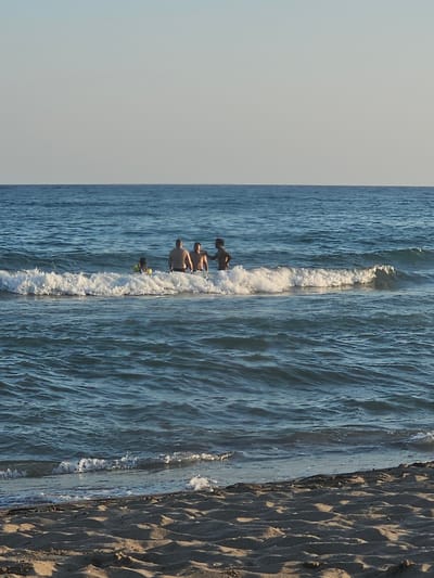 This image depicts a serene beach scene in Castelldefels, Spain, likely during the late afternoon given the soft, warm lighting casting long shadows on the sand. The composition is dominated by the meeting of the ocean and the shore, with the water occupying the majority of the frame. Gentle waves are rolling in, creating a foamy white line where they break.

In the water, four individuals are visible. The closest to the shore is a person in the water, seemingly engaging with the waves. Further out, three other individuals are also in the water, appearing to be enjoying the ocean. Their attire suggests casual beachwear, and their presence adds a sense of activity and enjoyment to the tranquil setting.

The sand on the right side of the image is textured, showing the distinct patterns left by the receding tide. The vast expanse of the ocean stretches towards the horizon, meeting a pale, cloudless sky. The overall mood is one of peaceful recreation and enjoyment of a summer's day. No text is visible in the image.
