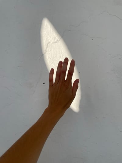 The scene, located in Varvarovka, Russia, depicts a human left hand and forearm extended diagonally upwards from the lower left towards the upper right. The hand is positioned against a white, textured wall, characterized by minor surface imperfections and hairline cracks. A bright, elongated, and irregular patch of direct sunlight illuminates a portion of the wall, extending vertically from the top-center downwards. The fingers of the hand are partially within this sunlit area, with the palm and lower arm in shadow. No other subjects or objects are discernible.