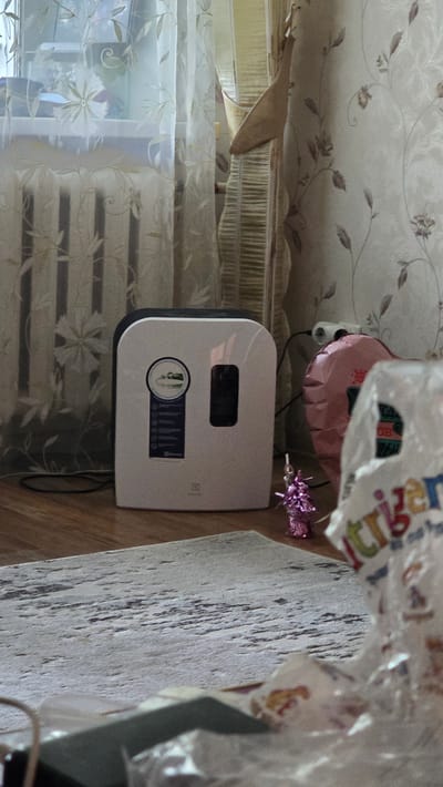 The image depicts an indoor scene, likely a living room, with a focus on a white and black air purifier positioned on a wooden floor. The air purifier has a circular sticker on its front with text and graphics, and a dark rectangular display panel. To the right of the air purifier, a pink heart-shaped balloon is visible, tethered by a string. In the foreground, a portion of a patterned rug is visible, along with a blurry object covered in clear plastic, which appears to be a gift with colorful text that partially reads "Tribute." The background features sheer curtains with a floral pattern drawn across a window and radiator. The overall lighting is dim, suggesting an indoor environment. The location is identified as Ukhta, Russia.