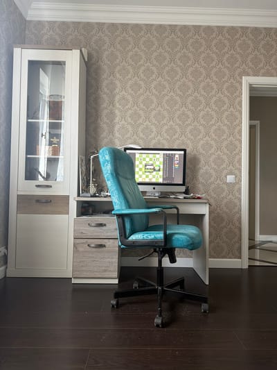This is a full-shot interior photograph of a home office, likely located in the village of Samakhvalavichy Rural Council, Belarus, due to the visual style consistent with residences in that region. The scene is centered around a desk with a computer and an office chair, suggesting a workspace.
The room features a patterned wallpaper in a neutral tone, a white crown molding along the top of the walls, and dark wood flooring. To the left of the desk is a tall cabinet with glass doors on the upper section, displaying various items, including what appears to be a bottle of Hennessy XO. The lower part of the cabinet has drawers.
The desk itself is white with a light wood-toned drawer section. On the desk, a computer monitor displays a chess game interface. Next to the monitor are a pair of glasses, some papers, and a pencil holder. A decorative clock and a few other small objects are also visible on the desk.
The focal point of the image is a bright teal-colored office chair with a black base and wheels, positioned directly in front of the desk. The chair appears to be made of a textured material, possibly leather or faux leather.
To the right of the desk, a doorway leads to another room, partially visible, with light-colored walls and a patterned floor. The overall impression is one of a functional and somewhat decorated home office.