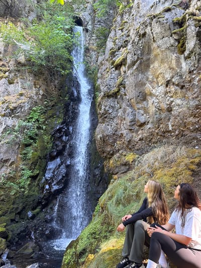 This image depicts Starvation Creek Falls, located in Wyeth, United States. Two adult females are seated on a moss and grass-covered bank situated to the right of the waterfall. Both individuals are looking upwards and to the left, towards the cascading water. The female on the left wears a black long-sleeved top, olive-green pants, and dark footwear. The female on the right wears a white short-sleeved t-shirt, black leggings, and white socks or low-profile shoes. The waterfall itself descends from a dark, circular culvert opening visible at the top of a tall, rugged rock cliff. The cliff face is characterized by exposed rock formations, dark wet sections, and patches of green moss and ferns. Water from the falls collects in a dark pool at the base before flowing into a visible stream in the foreground. Sparse green vegetation and larger trees are visible at the top and along the left side of the cliff. The scene is illuminated by natural daylight.