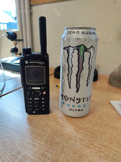 In a detailed view from Obruchishte, Bulgaria, a still life image depicts two primary objects resting on a light brown, wood-patterned surface, likely a desk. On the left, a black Motorola MTP850 professional radio stands upright. It features an extended black antenna, a dark screen displaying the Motorola logo, and a complete keypad with numerical and function buttons below the screen. To the immediate right of the radio, a white Monster Energy Ultra energy drink can is positioned vertically. The can is unopened and displays the distinctive black Monster claw logo, "MONSTER ENERGY ULTRA" text, and "ZERO SUGAR" prominently near its top rim. The background is softly out of focus, revealing a bright window with vertical structural elements on the left. A plain white wall is visible in the upper right background. Other indistinct items in the blurred background include dark cables, a black electronic device or stand, a yellow and blue writing instrument (possibly a pen) partially visible between the radio and the can, and a piece of white paper or card on the far right of the surface.