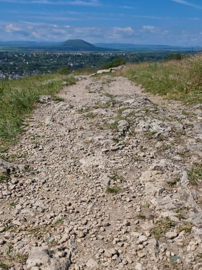 The image captures a rocky, upward sloping path leading towards a town nestled in a valley, with a distinctive conical mountain in the background. The path is covered with small stones, gravel, and sparse patches of grass. On either side of the path, longer grass grows wild. In the distance, a town with scattered buildings and trees is visible, surrounded by rolling green hills. The sky above is blue with scattered white clouds. The scene is set in Goryachevodsky, Russia, likely from an elevated vantage point overlooking the town and its surrounding landscape. No people are visible in the image.