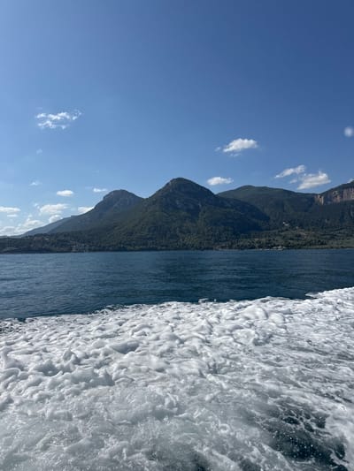 A high-angle, medium shot captures a scenic view of a body of water, with the Municipal Unit of Ayios Konstantinos, Greece, in the background. In the foreground, white foam churns from the movement of a boat, with the dark blue water stretching towards the shore. Behind the water, lush green mountains rise dramatically, their slopes covered in dense trees. The sky above is a clear blue, punctuated by a few scattered white clouds. The sun appears to be high in the sky, casting a bright light on the landscape. There are no people or animals visible in the image. The scene conveys a sense of natural beauty and tranquility.
