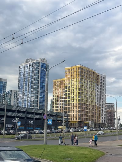 The image captures a street scene in the city of Senitsa, Belarus, featuring modern high-rise buildings and a busy road. In the foreground, a black car is partially visible on the left, with a grassy area and a sidewalk extending to the right. Several people are on the sidewalk, including a woman with a backpack, a group of adults and children, and a person on a scooter. A young child is also present. Across the road, a multi-story parking garage is visible with the signage "Салона" and "8-23". Above the parking garage, several contemporary apartment buildings dominate the skyline, with one prominent blue-windowed tower and a yellow and beige multicolored tower. Further in the distance, more apartment buildings can be seen. Overhead, power lines stretch across the cloudy sky. The environment appears to be urban, with evident infrastructure for transportation and housing.