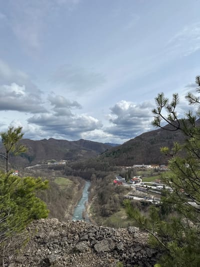 The image captures a scenic view from an elevated position in the city of Srednogortsi, Bulgaria. The foreground features rocky terrain and sparse vegetation, including coniferous trees, suggesting a mountainous or cliffside location.
In the midground, a river winds through a valley, flanked by forested hillsides and the built environment of the town. Buildings are visible along the riverbanks and further up the slopes.
The background reveals a mountainous range under a sky filled with dynamic cloud formations. The overall impression is one of a natural landscape in close proximity to human development.